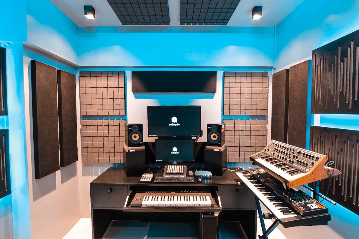 Production class: a desk with a monitor, keyboard controller, studio speakers and a synthesizer in a blue-lit treated room